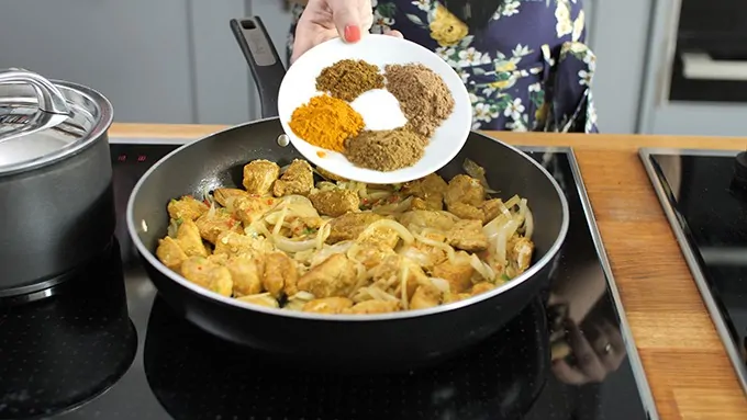 spices being added to a pan with chicken and onions