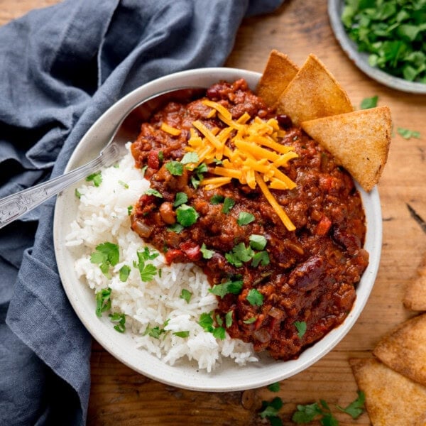 A bowl of chilli con carne with kidney beans, ground beef, cheese, and cilantro over white rice. Tortilla chips on the side.