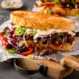 Close-up of a loaded Korean steak sandwich with melted cheese, sliced red onions, jalapeños, and peppers on toasted bread, served on a sheet of paper with a knife.
