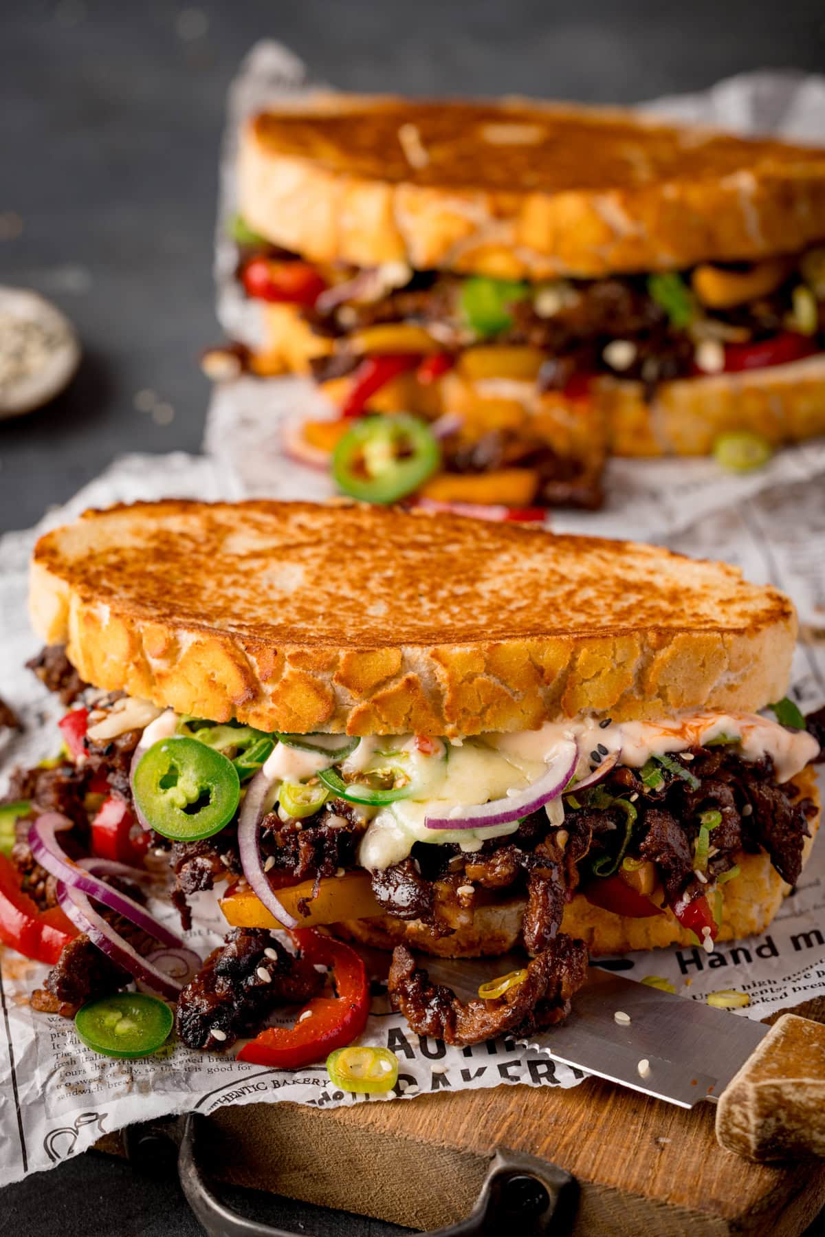 Two toasted Korean steak sandwiches with bulgogi, jalapeños, sliced red onions, fried peppers, and garlic mayo on a wooden board lined with newspaper.