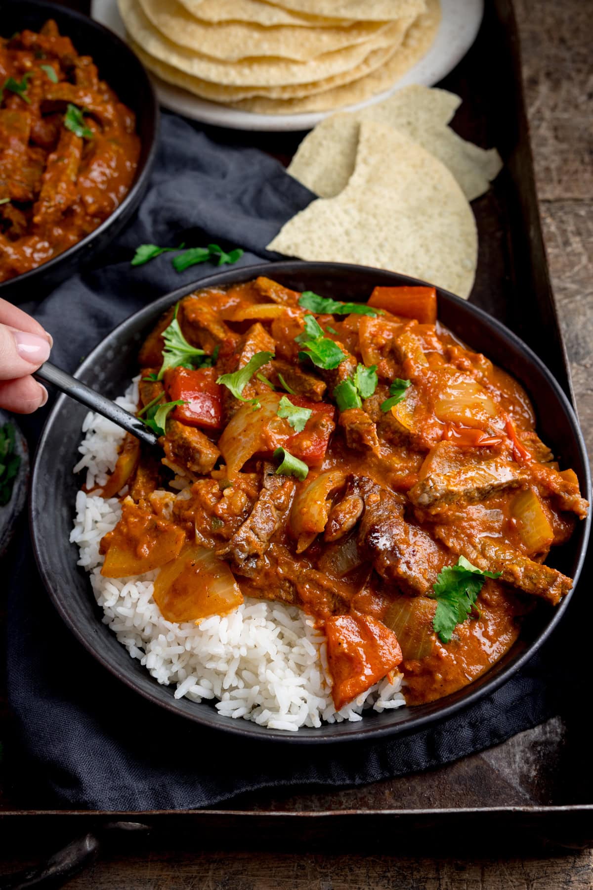A dark bowl with boiled rice and steak curry, garnished with coriander (cilantro) on a dark tray. A forkful of the curry is being taken. There is a further bowl of curry and broken poppadoms in the background.